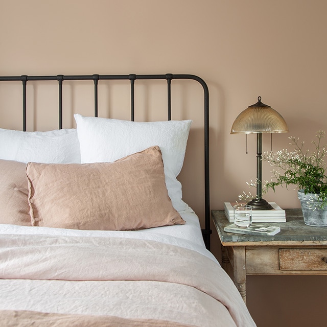 A wrought-iron bedframe with soft neutral-colored bedding pairs beautifully with a warm light clay painted wall and a rustic side table with a lamp. A wrought-iron bedframe with soft neutral-colored bedding pairs beautifully with a warm light clay painted wall and a rustic side table with a lamp.