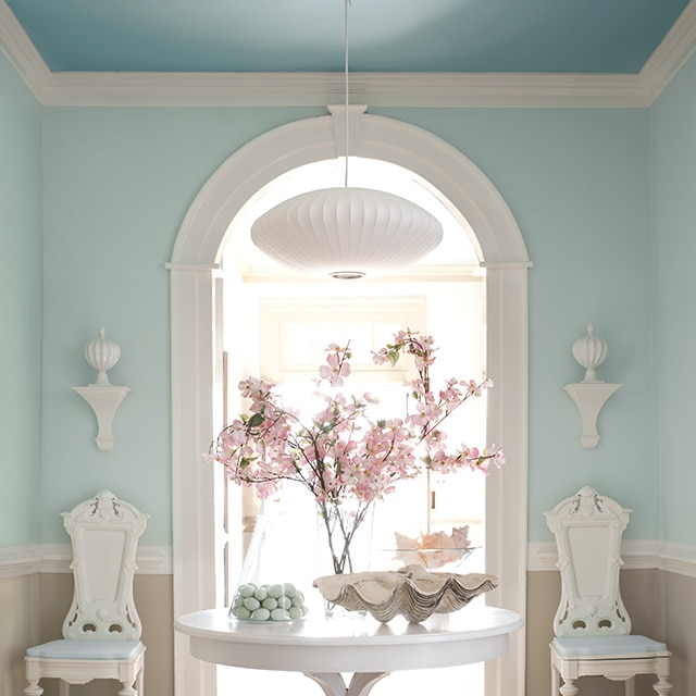 A spacious hallway with light blue painted upper walls, neutral lower walls, and a blue ceiling features a white table and chairs, and a white painted arch and trim throughout. A spacious hallway with light blue painted upper walls, neutral lower walls, and a blue ceiling features a white table and chairs, and a white painted arch and trim throughout.