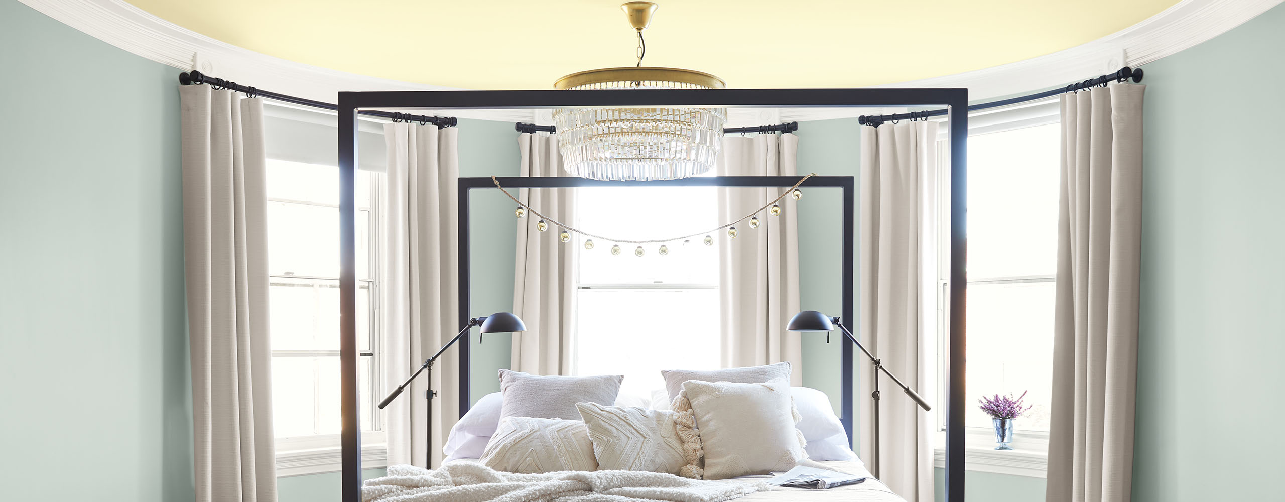 Rounded bedroom walls painted a soft blue-gray, a pale yellow ceiling, white trim, and three windows surround a comfy black four-poster bed. Rounded bedroom walls painted a soft blue-gray, a pale yellow ceiling, white trim, and three windows surround a comfy black four-poster bed.