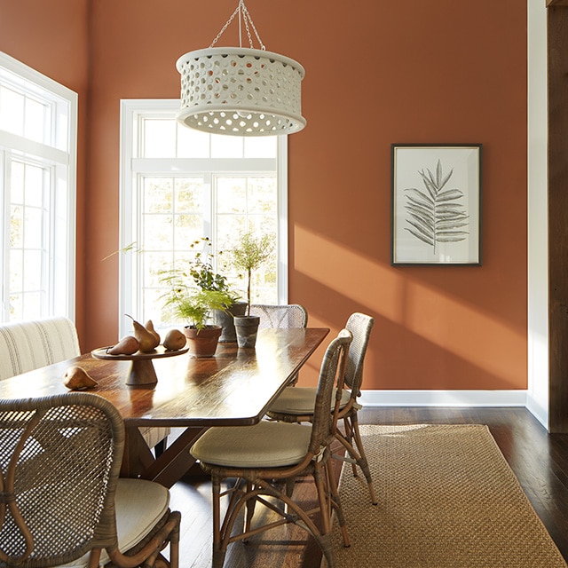 A sunlit dining room with deep burnt sienna painted walls, simple framed art, and a wood table with rattan chairs. A sunlit dining room with deep burnt sienna painted walls, simple framed art, and a wood table with rattan chairs.