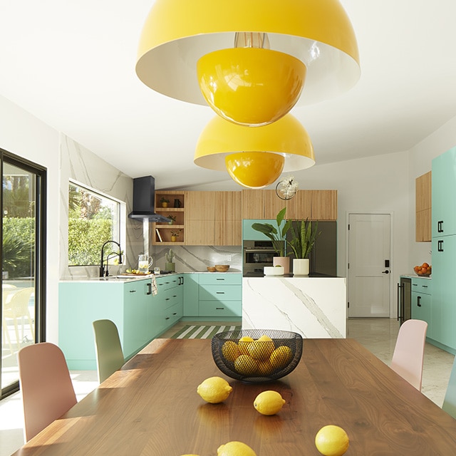 A cheerful dining area with a wood table, pastel-coloured chairs, and yellow hanging lamps next to an open kitchen with white walls and aquamarine painted cabinets. A cheerful dining area with a wood table, pastel-coloured chairs, and yellow hanging lamps next to an open kitchen with white walls and aquamarine painted cabinets.