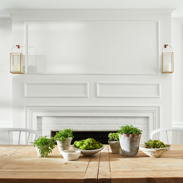 An inviting dining room with white painted walls, wall mantel, and ceiling, and white chairs behind a wood table topped with plants. An inviting dining room with white painted walls, wall mantel, and ceiling, and white chairs behind a wood table topped with plants.