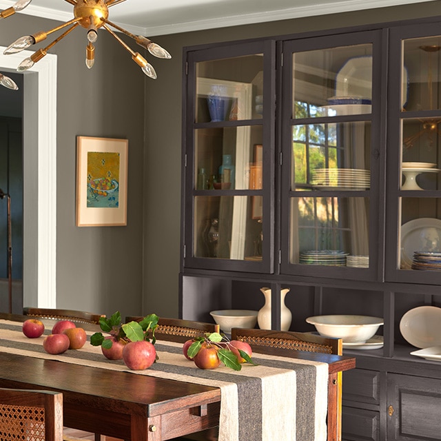 A gray painted dining room with white trim, a black cabinet displaying dinnerware, a wooden table with a striped runner, and caned chairs. A gray painted dining room with white trim, a black cabinet displaying dinnerware, a wooden table with a striped runner, and caned chairs.