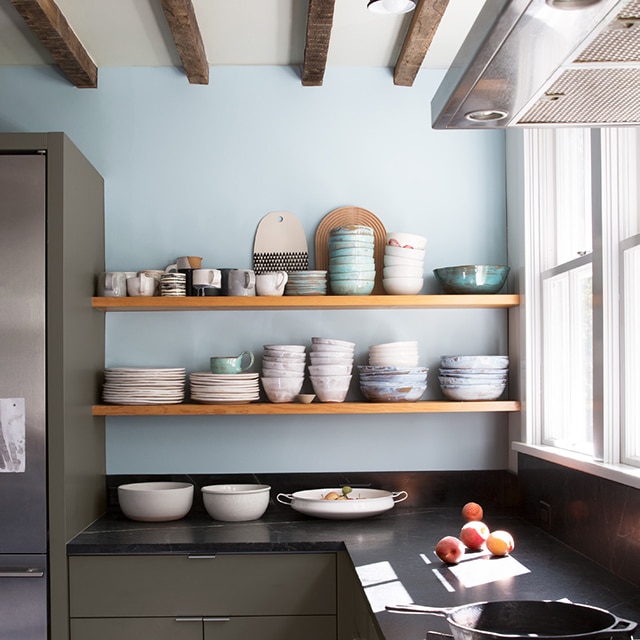 A rustic kitchen with a light blue-painted accent wall, dark mossy green-painted lower cabinets, open wooden shelving piled with bowls and plates, and a black countertop.