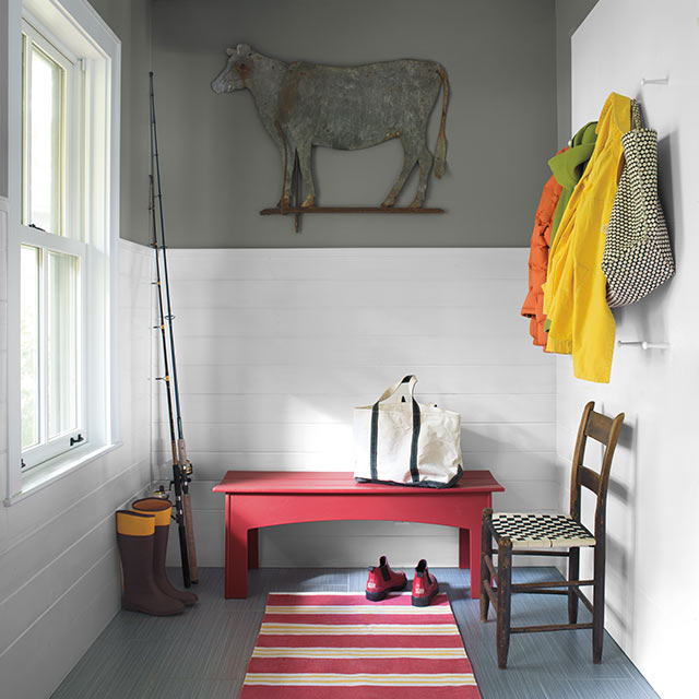 A mudroom with white wainscoting halfway up a grey wall featuring a red bench, red and white striped rug, and jackets hung on the wall.
