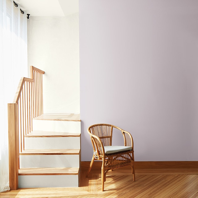 A small staircase with wood steps and railing, and white painted risers in the corner of a sunlit room with a white wall and ceiling, a light purple accent wall, and rattan chair. A small staircase with wood steps and railing, and white painted risers in the corner of a sunlit room with a white wall and ceiling, a light purple accent wall, and rattan chair.