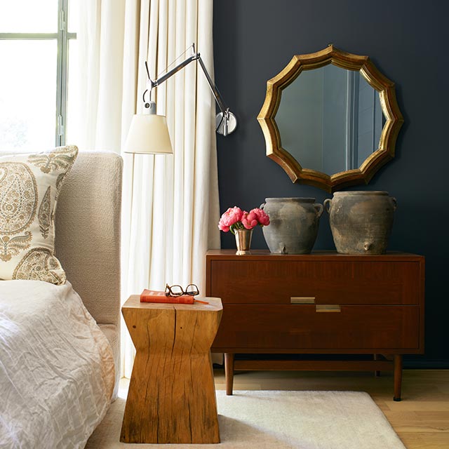 Une chambre à coucher peinte en noir arborant un lit crème, des tables d’accent en bois, des pots et des fleurs, un miroir doré et des rideaux blancs.