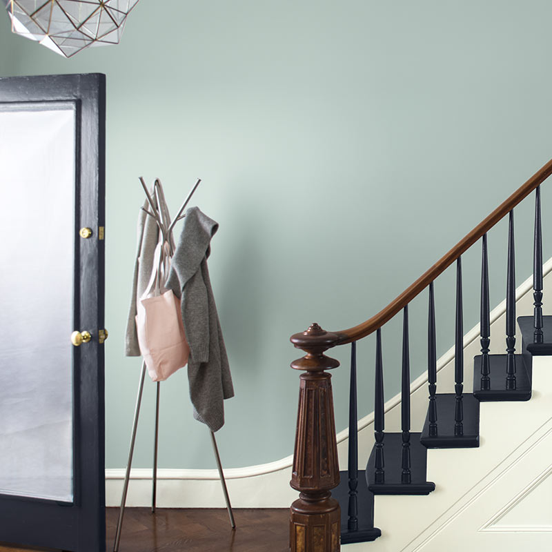 A blue-gray wall with white trim sets off a staircase with black stairs and an ornate wood banister. Color of the Year 2019