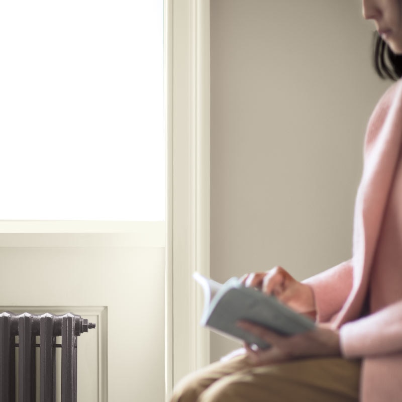 A woman sits reading next to a window with white trim and gray-beige-painted wall. Color of the Year 2019