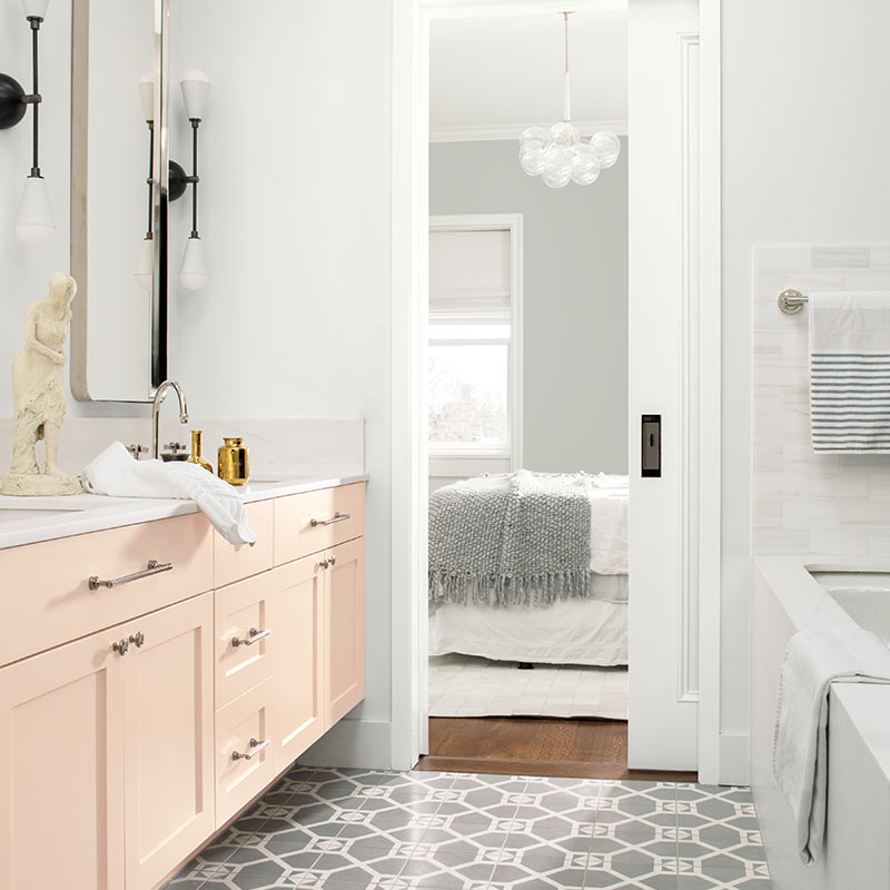 A white-painted bathroom with pale pink cabinets opens to a gray bedroom with contemporary chandelier. Color of the Year 2019