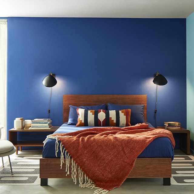 A contemporary bedroom with a dark blue painted wall, a light blue painted wall, a white ceiling, and a wooden bedframe and rust-colored chenille throw.