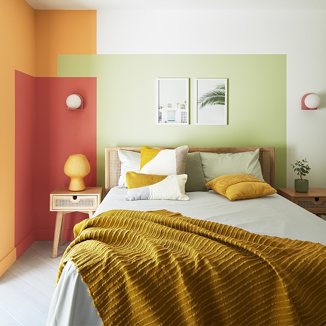 A small, contemporary bedroom with a painted color block wall design in green, red and orange against white-painted walls, and a double bed with an orange throw.