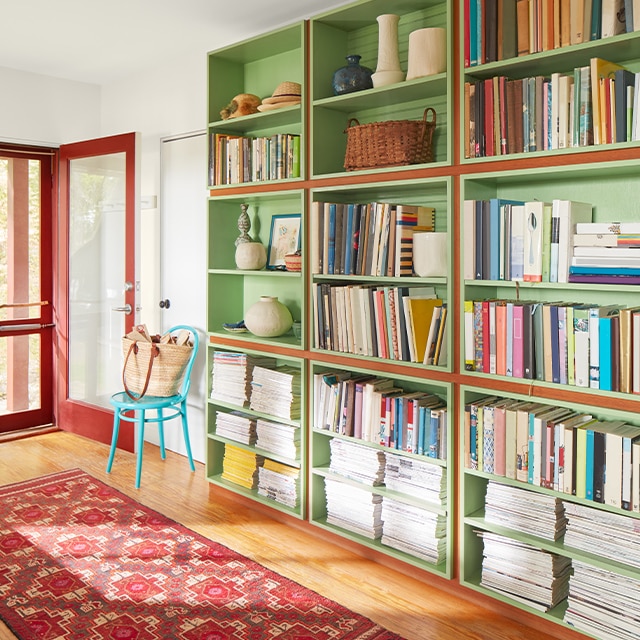 A sunlight entryway with green-painted bookshelves, wood floors, a red-painted front door, and a red patterned rug.
