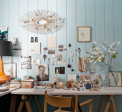 An eclectic selection of mementos, art and reminders are pinned against a light blue, wood-panelled wall over a sawhorse table.