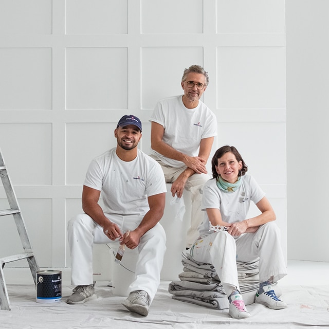 Three Benjamin Moore painting contractors dressed in white, posing in a white-painted room.