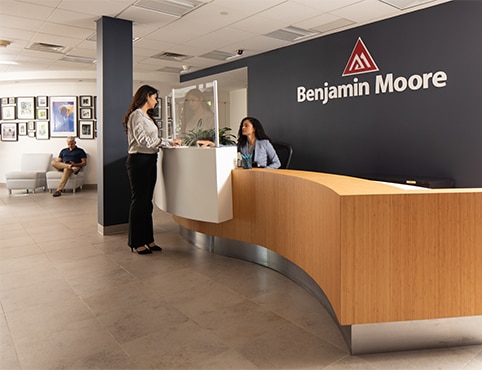 A person seated behind a long, wooden reception desk in the Benjamin Moore Montvale office greets another individual, while another person sits in the waiting area.