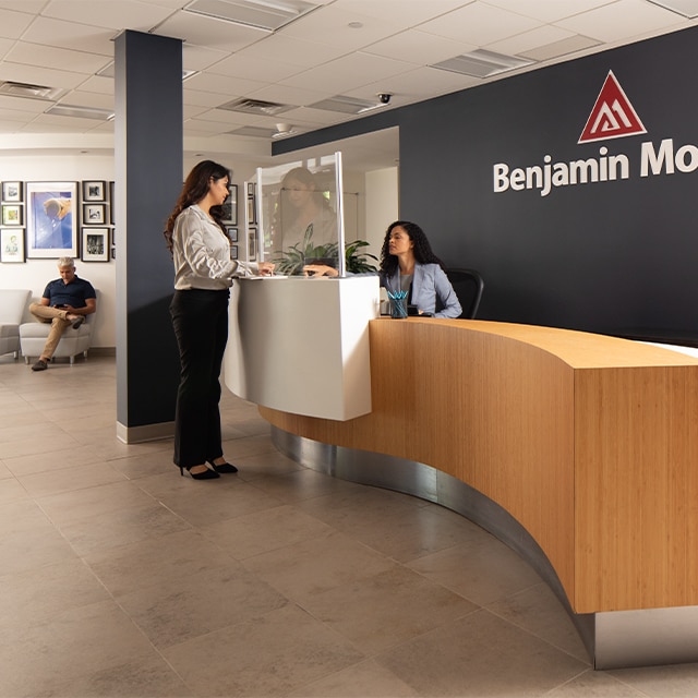 A person seated behind a long, wooden reception desk in the Benjamin Moore Montvale office greets another individual, while another person sits in the waiting area.