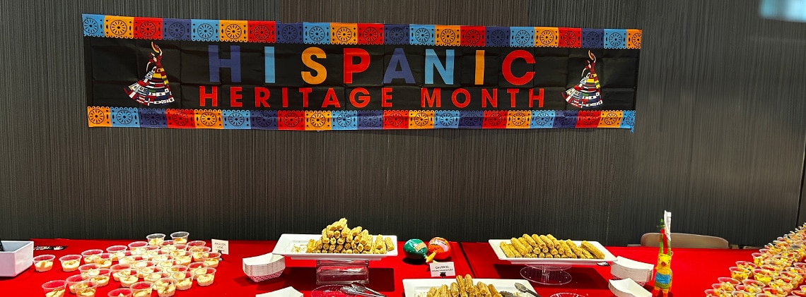 A colorful Hispanic Heritage Month banner hangs on a wall behind a red table displayed with food.