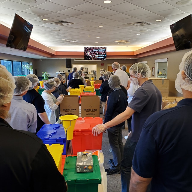 A group of Benjamin Moore employee volunteers with colored plastic containers watching an instructional video on how to safely pack food kits.