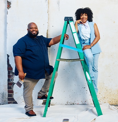 Two people leaning against a green ladder, pose in front of a white wall with exposed brick in need of paint and repair.