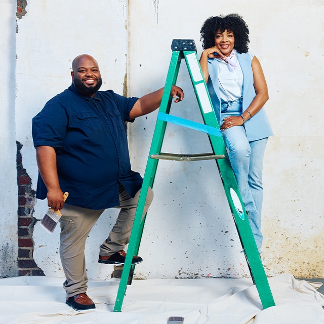 Two people leaning against a green ladder, pose in front of a white wall with exposed brick in need of paint and repair.