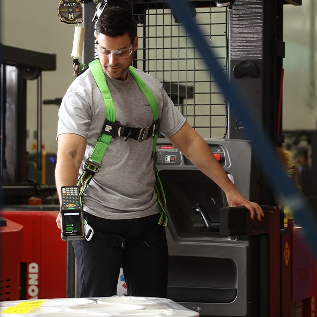 A Benjamin Moore employee wearing a green harness and safety glasses stands next to heavy equipment while holding a scanner over white shrink wrapped containers.