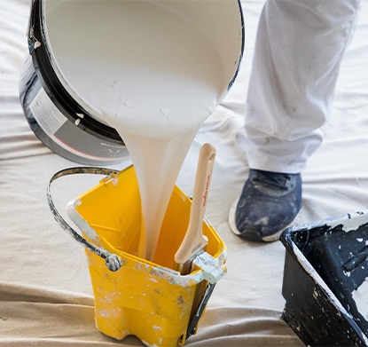 A bucket of white paint being poured into a smaller yellow bucket on top of a white drop cloth.
