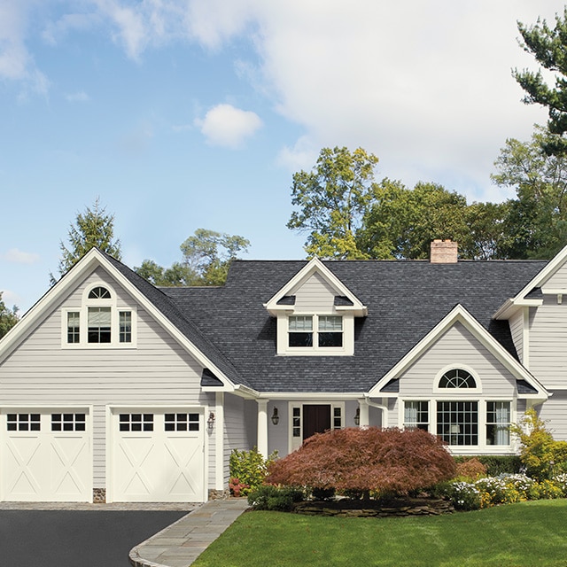 A pretty light gray-painted home exterior with white garage doors and trim, a lush green lawn and landscaping.