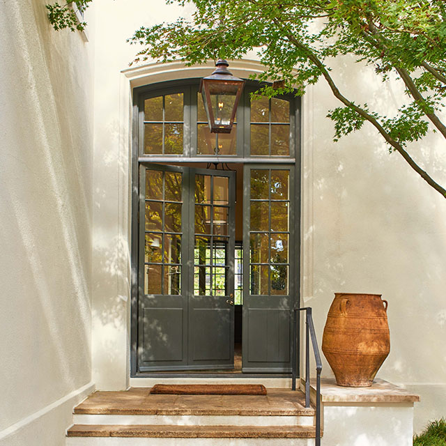 An off-white painted stucco house with an elegant dark green front door with glass panes and transom windows, ceramic tile steps and landing with a large clay pot, partially shaded by a tree.