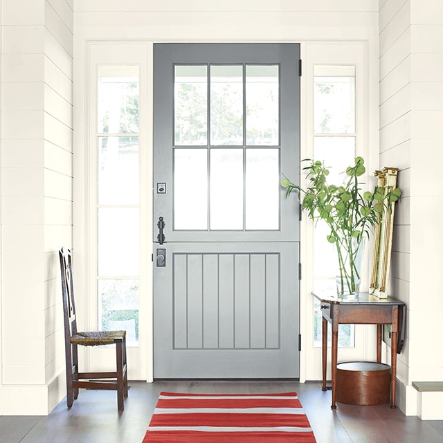 A bright and welcoming home entryway with a gray painted front door, white shiplap walls, a wood table and chair, and red-and-gray striped rug.