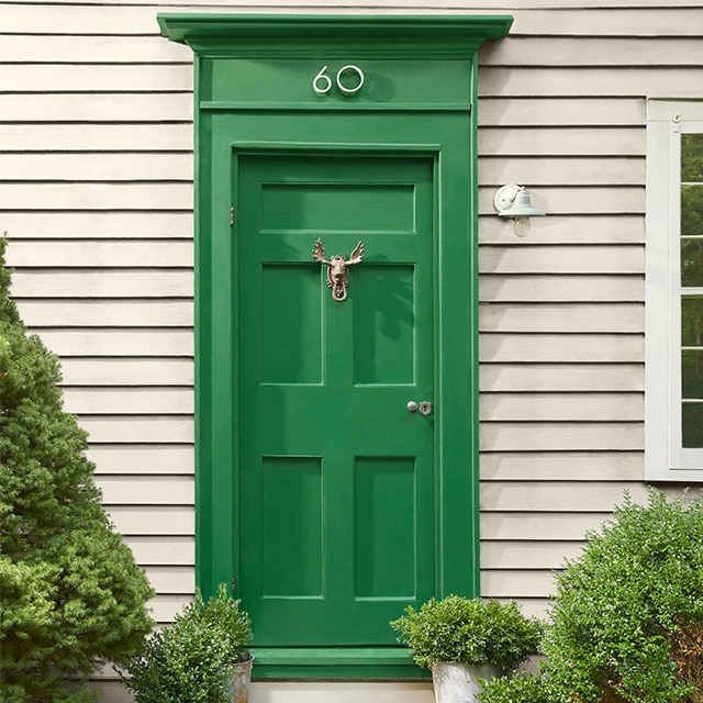 Warm greige-painted house siding, with a pretty green door flanked by shrubs, and white window trim.