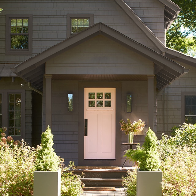 A soothing pink-painted front door makes a lovely contrast on this soft black-painted house, nestled among trees and front shrubs.