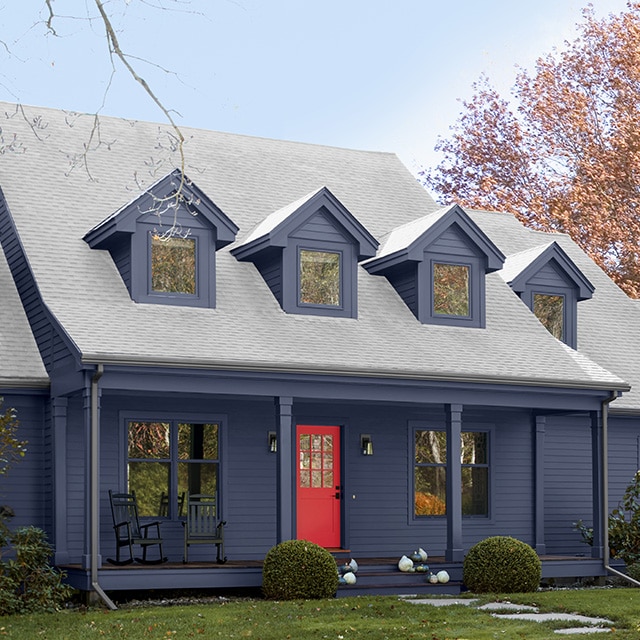 A beautiful navy blue painted Cape Cod style home features dormer windows, a front porch with pillars, and a striking red painted front door.