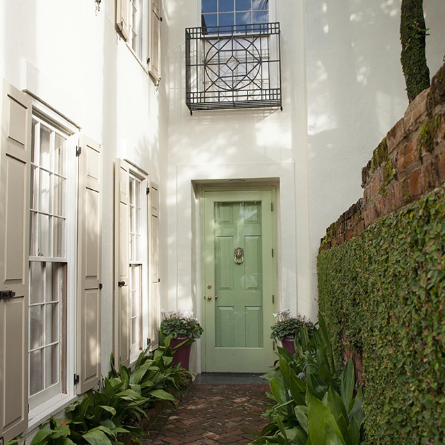 The front entrance of an elegant, white-painted house with beige shutters, a light-green front door, brick walkway and brick wall covered in foliage.