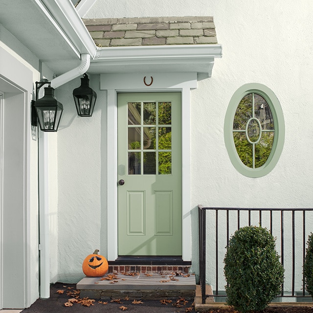 The side entrance of a white painted stucco home with a light-green painted door and oval-framed window, two black hanging lanterns, and a pumpkin on the doorstep.