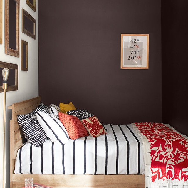 Una habitación acogedora con paredes en matices chocolate intenso, una pared de galería pintada de blanco, una cama con ropa de cama a rayas blancas y negras, almohadones y una manta con estampado rojo, y una otomana de mimbre.