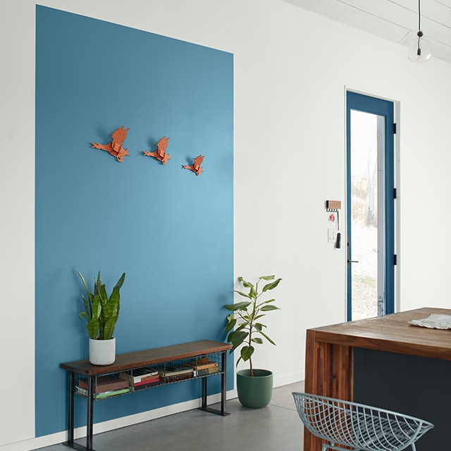 Una entrada contemporánea pintada de blanco con una puerta con marcos azules y un gráfico rectangular pintado de azul intenso detrás de un banco de madera, y una isla de madera pintada de gris carboncillo con un taburete de bar de malla.