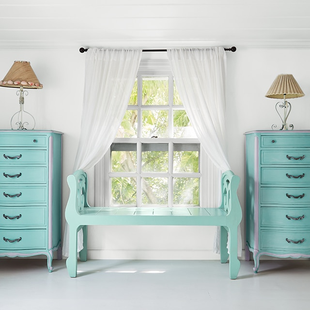 Una alegre habitación pintada de blanco con un toque de gris azulado, un banco turquesa y una ventana con cortinas blancas flanqueadas por cómodas pintadas de azul turquesa con una lámpara en cada una.