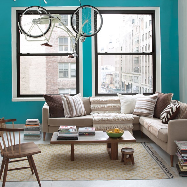Sala de estar luminosa de un apartamento urbano con una pared de acento pintada de color turquesa, paredes laterales y marcos blancos, dos ventanas grandes, muebles de color beige y marrón, y una bicicleta colgada de tuberías expuestas.