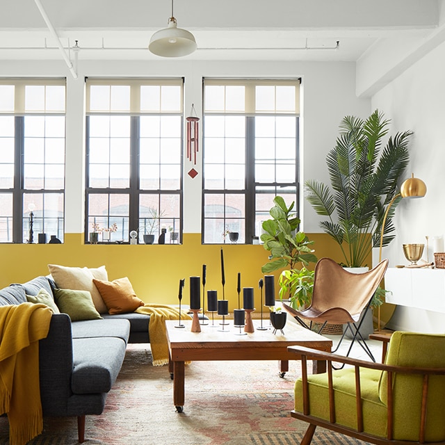 Una sala de estar liviana estilo loft con una pared y un techo pintados de blanco con tuberías expuestas, una pared de acento en blanco y amarillo con ventanas altas y muebles modernos en gris, amarillo y madera.
