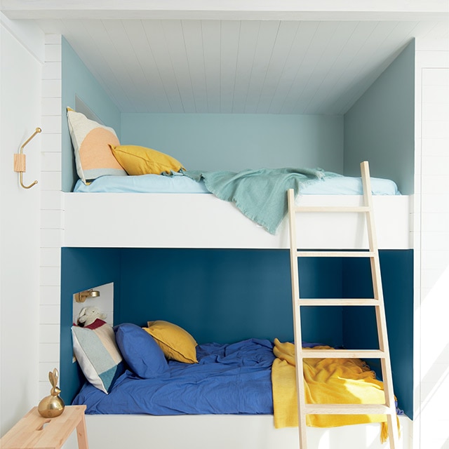 Una habitación pintada de color blanco con literas integradas en tonos azules con prendas de cama haciendo juego, almohadores amarillos, un taburete de madera y una escalera.