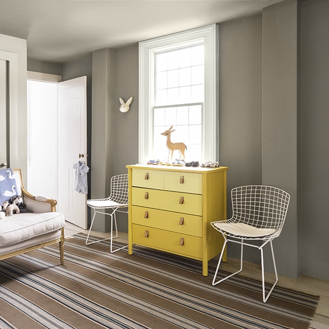 A room with gray painted walls, white trim and doors, a yellow ombré painted wood dresser flanked by two white metal chairs, and a beige sofa with stuffed animals.