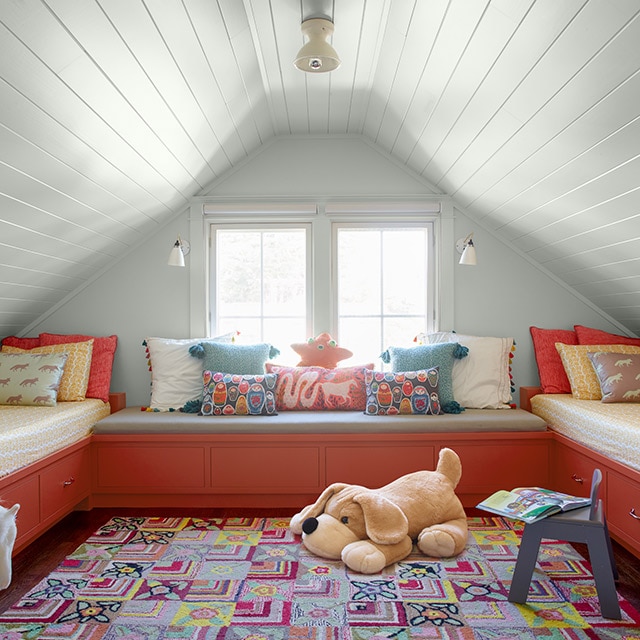 A white painted kids’ bedroom with a vaulted shiplap ceiling features colourful bedding and decor, a coral built-in window seat, and twin bed frames with drawers beneath. 