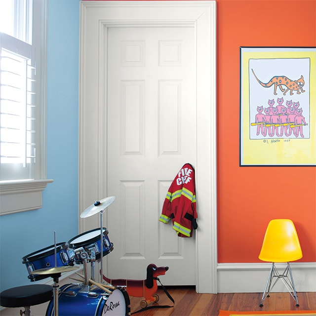 Una sala de juegos infantil con paredes coloridas en tonos naranja y azul, puerta y marcos en color blanco, una batería, arte enmarcado y una silla amarilla.