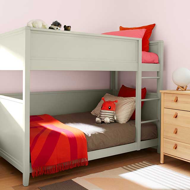 A soft pink painted kids’ bedroom features light jade green painted bunk beds with gray and red bedding, a wooden dresser, and a gray striped rug.