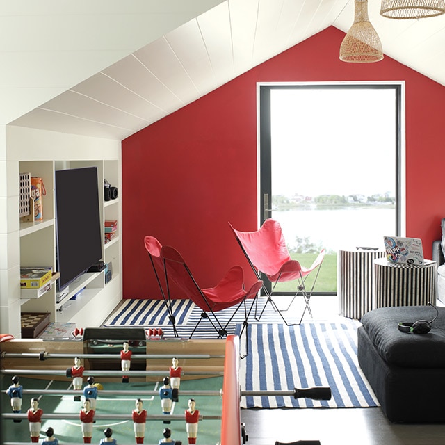 A white painted game room with built-in shelves and TV, a vaulted shiplap ceiling, a red accent wall with a glass door, billiard and foosball tables, a black sofa, and red chairs. 