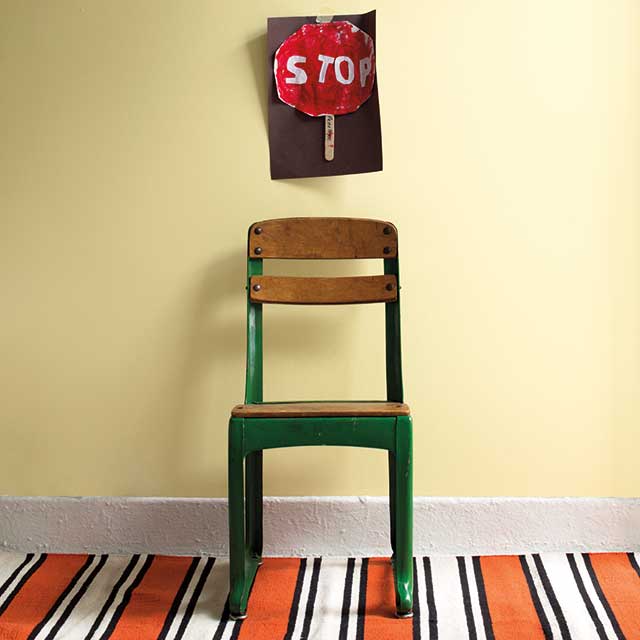 Una silla de escuela de madera verde contra una pared pintada de amarillo claro, con un cartel de pare pintado a mano en la pared, y una alfombra rayada de color naranja, blanco y negro.