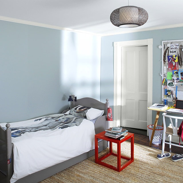 A kid’s bedroom with blue painted walls and white trim, ceiling, and door also features a gray bed with astronaut bedding, a red table, and a desk and chair under a bulletin board.