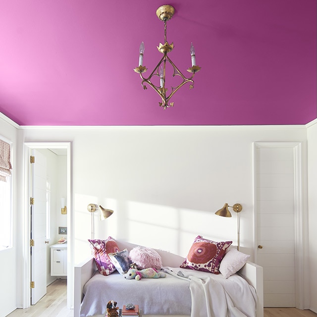 Un techo pintado en rosa oscuro añade un toque divertido de color a esta habitación blanca con alhomadones de múltiples colores y ropa de cama blanca sobre una cama blanca, ubicada entre dos puertas.