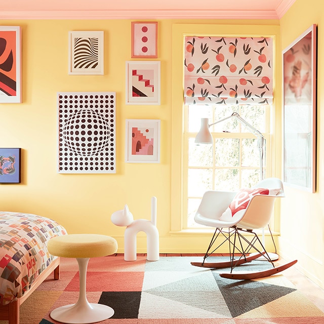 Una habitación de niño brillante y alegre con paredes pintadas en amarillo, un techo rosado pastel, arte con marcos coloridos, muebles, modernos, un cubrecamas de múltiples colores y una alfombra con estampado geométrico.